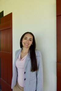 Woman with long dark hair, in a light blazer and pink top, smiles by red shutters—trusted personal injury lawyers Louisiana.
