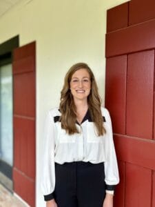 A woman with long brown hair stands indoors, dressed in a white blouse and black pants, near a red and white wall at immigration lawyers Louisiana.