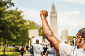 A group protests outdoors; one person holds a face mask and raises a fist. A tall building, home to immigration visa lawyers, looms behind them.
