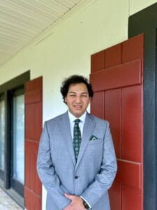 A person in a gray suit, possibly one of the top visa lawyers, stands smiling before a white wall with red window shutters.
