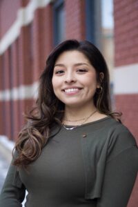 A woman with long brown hair in a green top and layered necklaces smiles, standing in front of a red brick wall near immigration lawyers Louisiana.