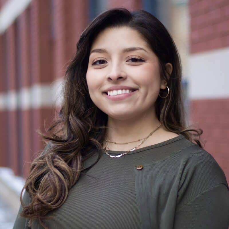 A woman with long brown hair in a green top and layered necklaces smiles, standing in front of a red brick wall near immigration lawyers Louisiana.