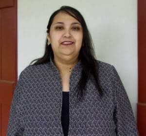A woman with long dark hair, possibly one of the top immigration lawyers Louisiana, stands by a white wall with a red door frame.