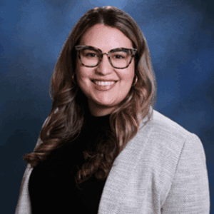 Woman with long wavy hair, glasses, and a light blazer over a black top, smiling like one of the top personal injury lawyers Louisiana trusts.