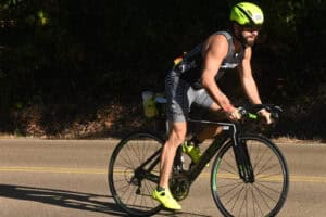 A man wearing a neon yellow helmet and shoes rides a black bicycle past a deportation law firm on a paved road in daylight.