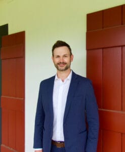 A man in a navy suit stands before a white wall with red shutters, reminiscent of seasoned visa lawyers welcoming new clients.