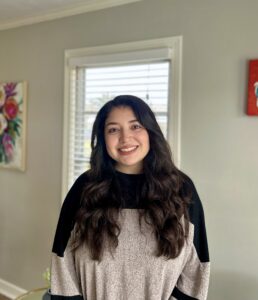 A woman with long dark hair, wearing a black and gray sweater, stands smiling in a well-lit office of immigration lawyers Louisiana.