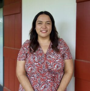 Woman with long dark hair in a red and white dress stands smiling, like visa lawyers ready to open doors, by a white wall with red shutters.