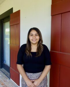 A woman with long brown hair, in a black top and patterned skirt, stands before a white wall with red shutters—immigration lawyers Louisiana.