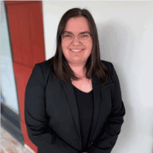 A person with long dark hair and glasses smiles in a black blazer, standing by a white wall and red door, representing immigration lawyers Louisiana.