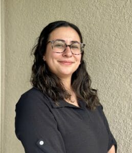 A woman with long dark hair and glasses, possibly one of Louisiana’s skilled immigration lawyers, smiles softly before a light-textured wall.