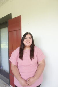 A person with long dark hair in a pink top stands by a white wall with red shutters, smiling—perhaps a visa lawyer in Louisiana.