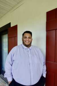 A man in a light purple button-up shirt stands before red window shutters, smiling—like a trusted criminal defense attorney in Louisiana.