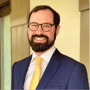 Man with glasses, beard, and mustache in a blue suit—one of the top personal injury lawyers Louisiana—smiling indoors at the camera.