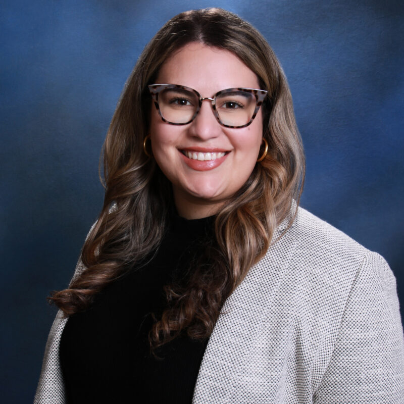 Smiling woman with long wavy hair, glasses, and a light-colored blazer—Nia Reichard poses in front of a blue gradient background.