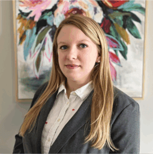 A woman with long blonde hair in a gray blazer stands before a colorful floral painting, representing immigration lawyers Louisiana.