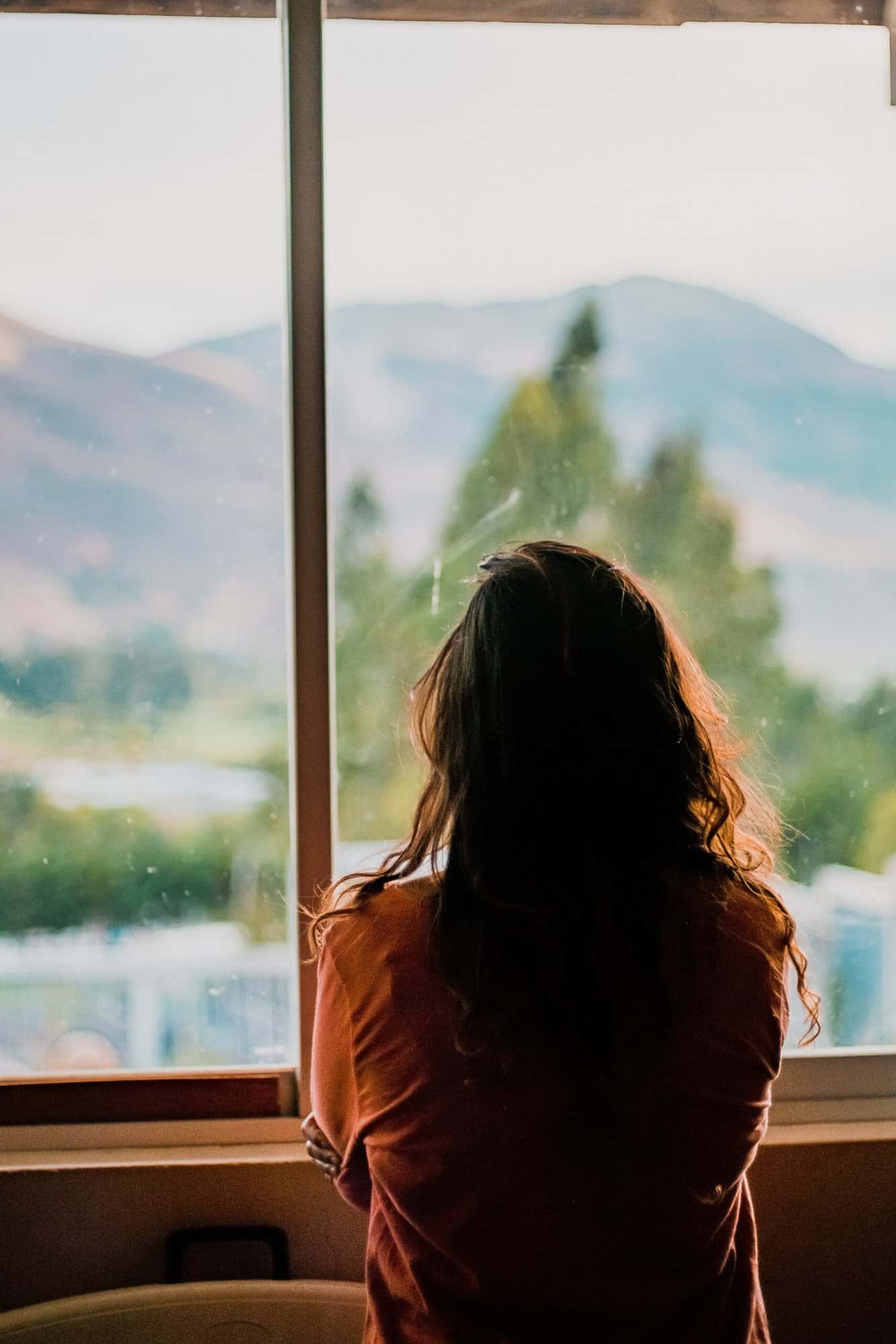 A person with long hair stands indoors, looking out a window, perhaps reflecting on advice from immigration visa lawyers.