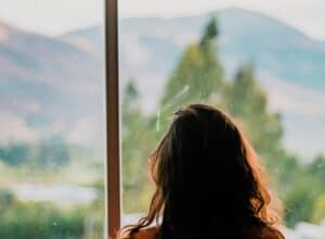 A person with wavy hair looks out a window, contemplating distant mountains—much like immigration visa lawyers planning new journeys.