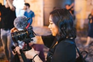 A woman holds a camera with a mic, filming outdoors at night, as people and personal injury lawyers Louisiana walk in the background.