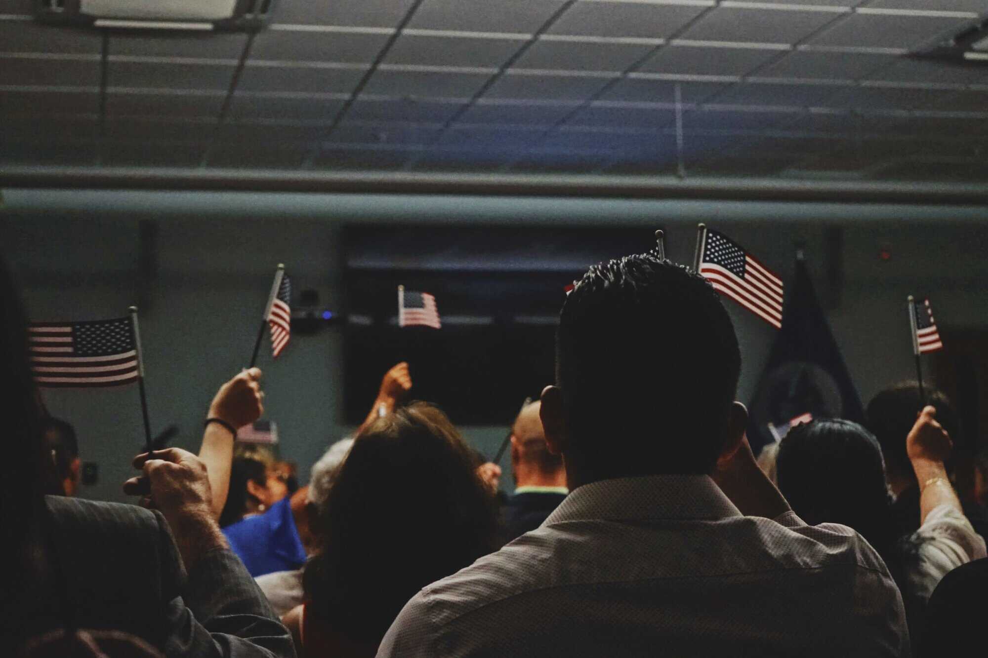 A group of people indoors holding small American flags, perhaps awaiting immigration visa lawyers, viewed from behind under fluorescent lights.
