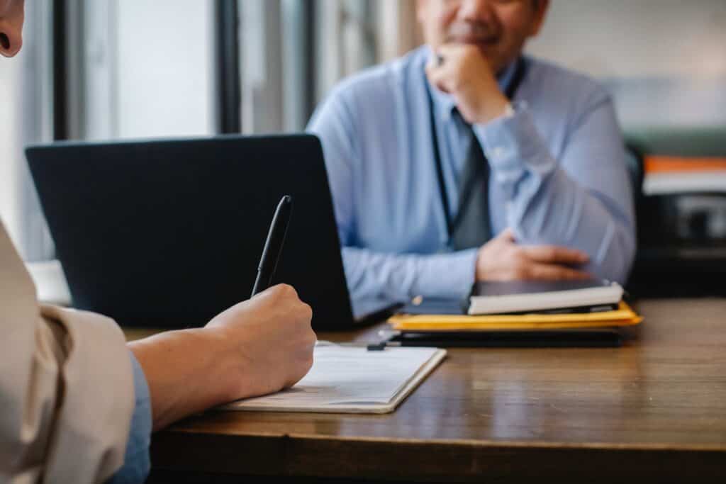 Two people sit at a desk with documents as a louisiana immigration lawyer writes on a clipboard while the other observes.