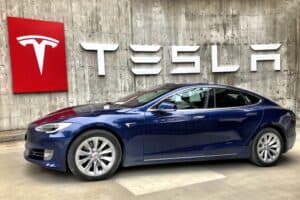 A blue Tesla sedan is parked before a concrete wall with a large Tesla logo and sign for immigration visa lawyers.