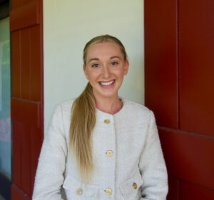 Kelsey Sulik, with long blonde hair in a ponytail, stands before a red and white wall, wearing a light-colored textured jacket.