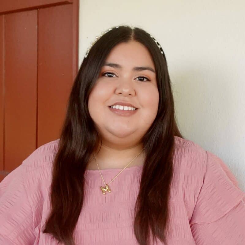 A woman with long dark hair wearing a pink top and gold necklace sits in front of a red door and off-white wall, smiling at the camera.