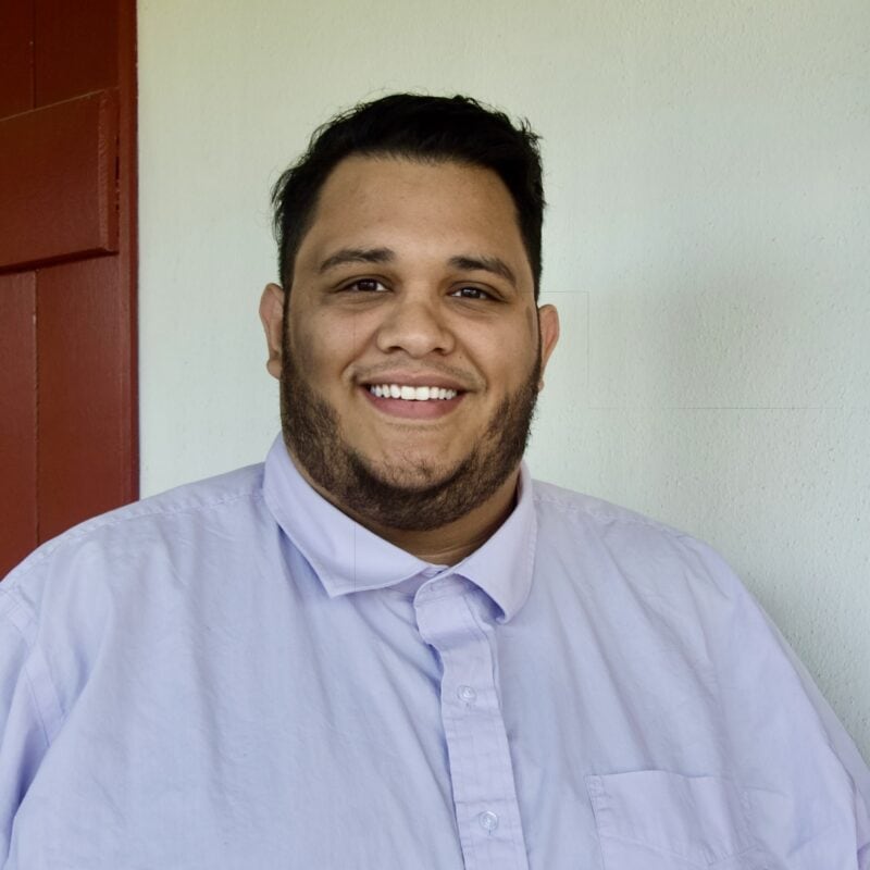 A man with short dark hair and a beard, wearing a light purple button-up shirt, stands smiling in front of a white wall and red door.