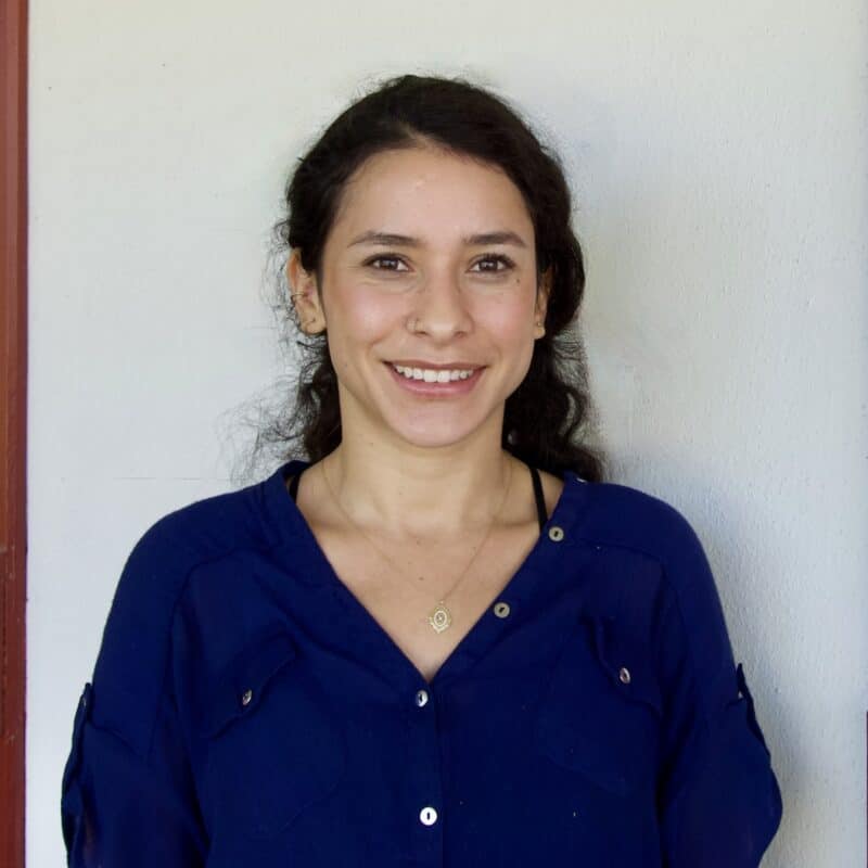 A woman with long dark hair and a blue blouse smiles at the camera in front of a plain off-white wall.