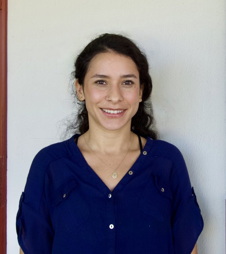 A woman with long dark hair and a blue blouse smiles at the camera in front of a plain off-white wall.