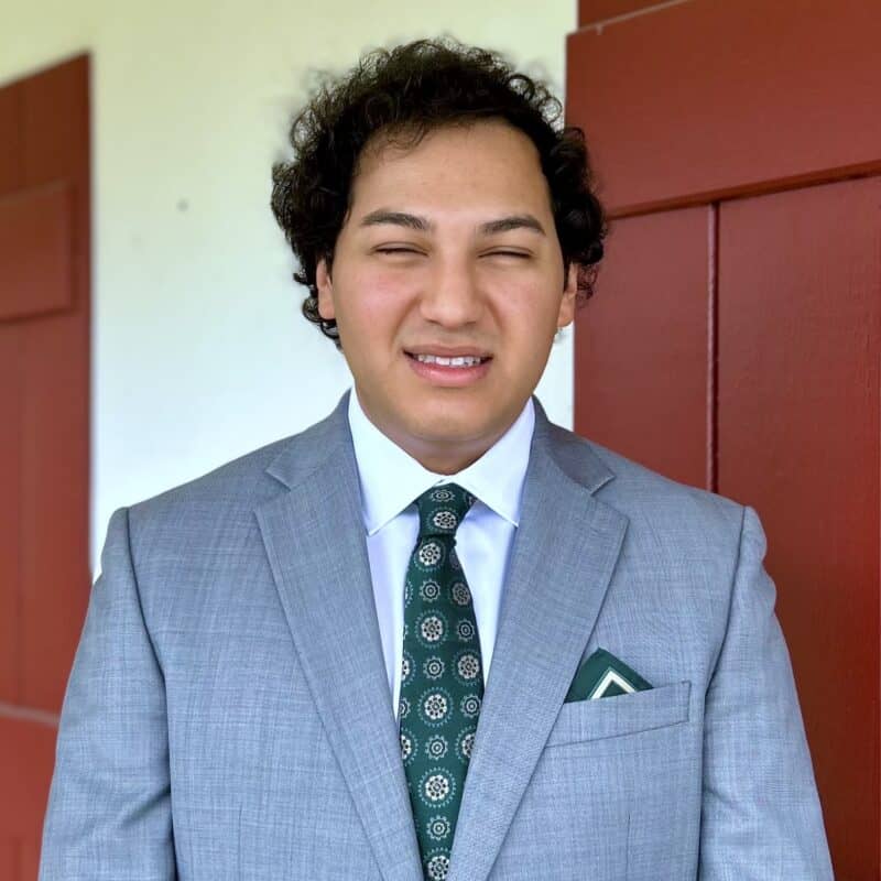 A person in a gray suit and patterned green tie stands in front of a red door, looking at the camera and smiling slightly.