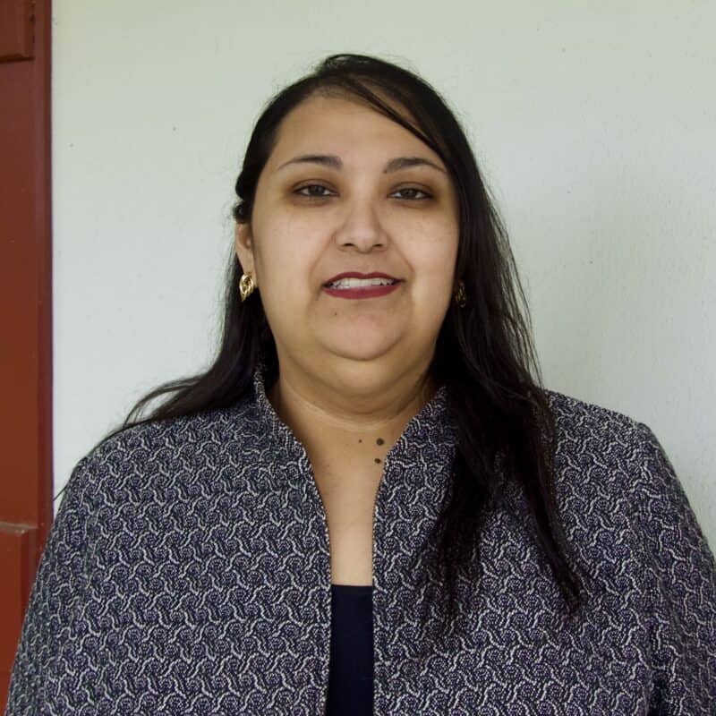 A woman with long dark hair wearing a patterned jacket stands in front of a light-colored wall and looks at the camera.