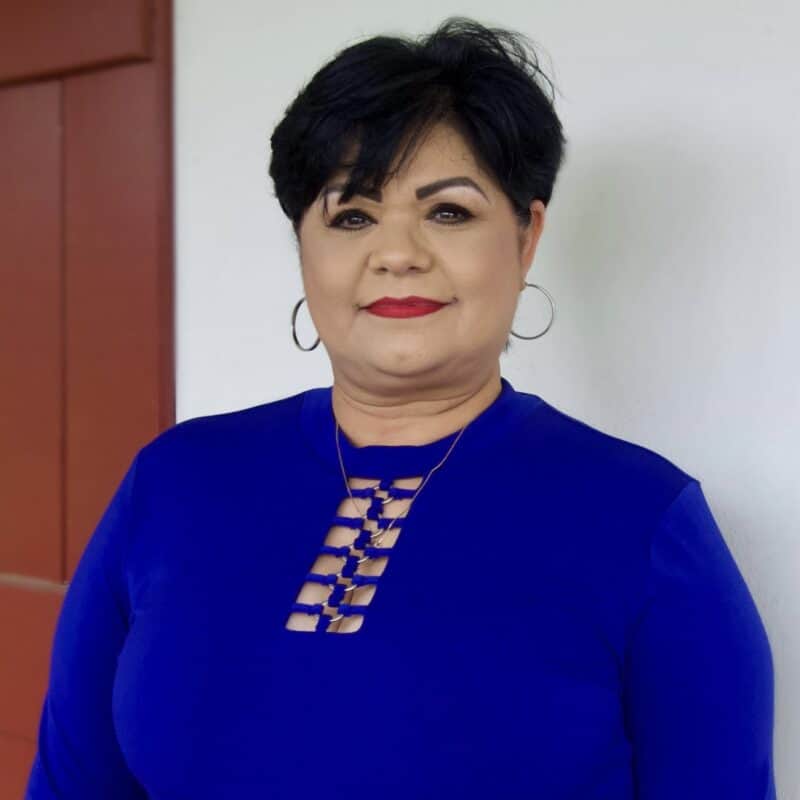 A woman with short dark hair and hoop earrings wears a blue top and stands in front of a white wall with a red door.