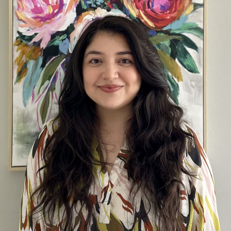 A woman with long dark hair stands in front of a colorful floral painting, wearing a patterned blouse and smiling at the camera.