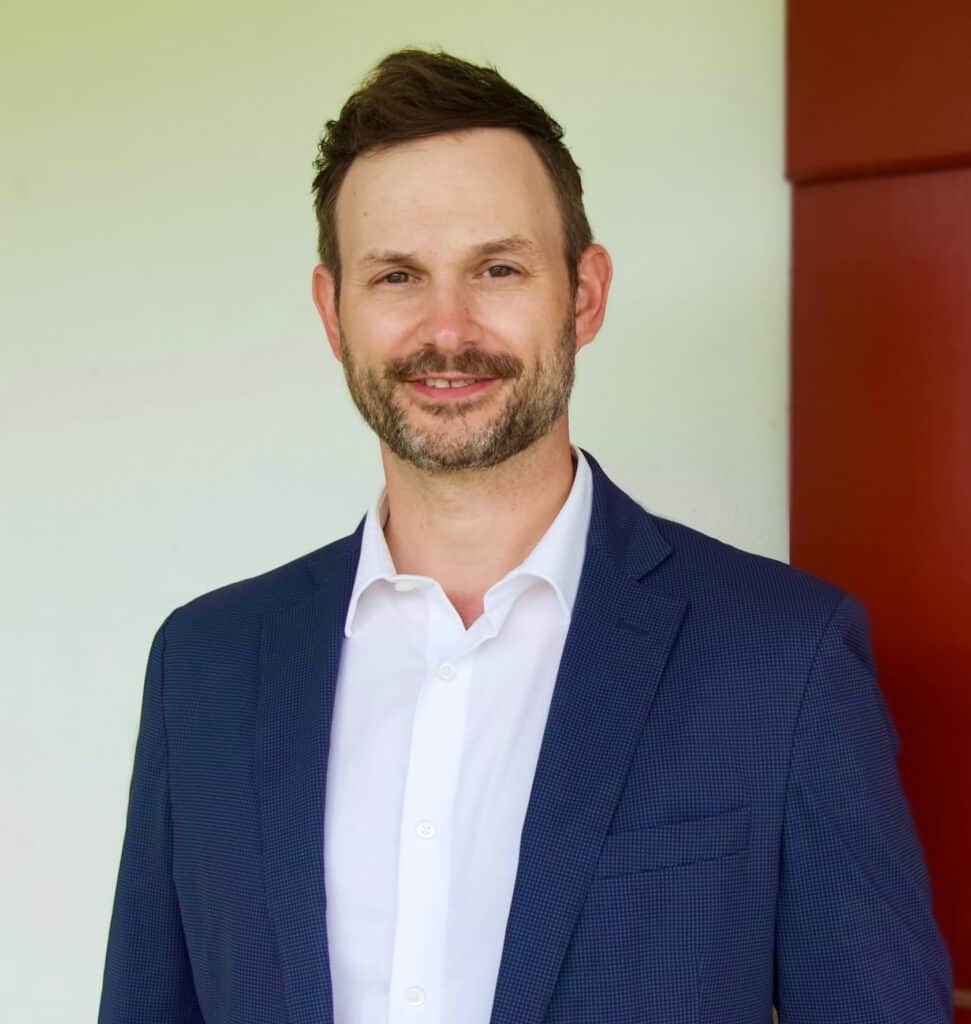 Man with short brown hair and beard wearing a navy suit jacket and white shirt, standing indoors against a light background.