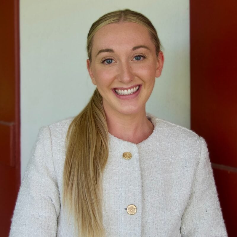 A woman with long blonde hair in a low ponytail, wearing a white textured jacket, smiles in front of a white and red background.