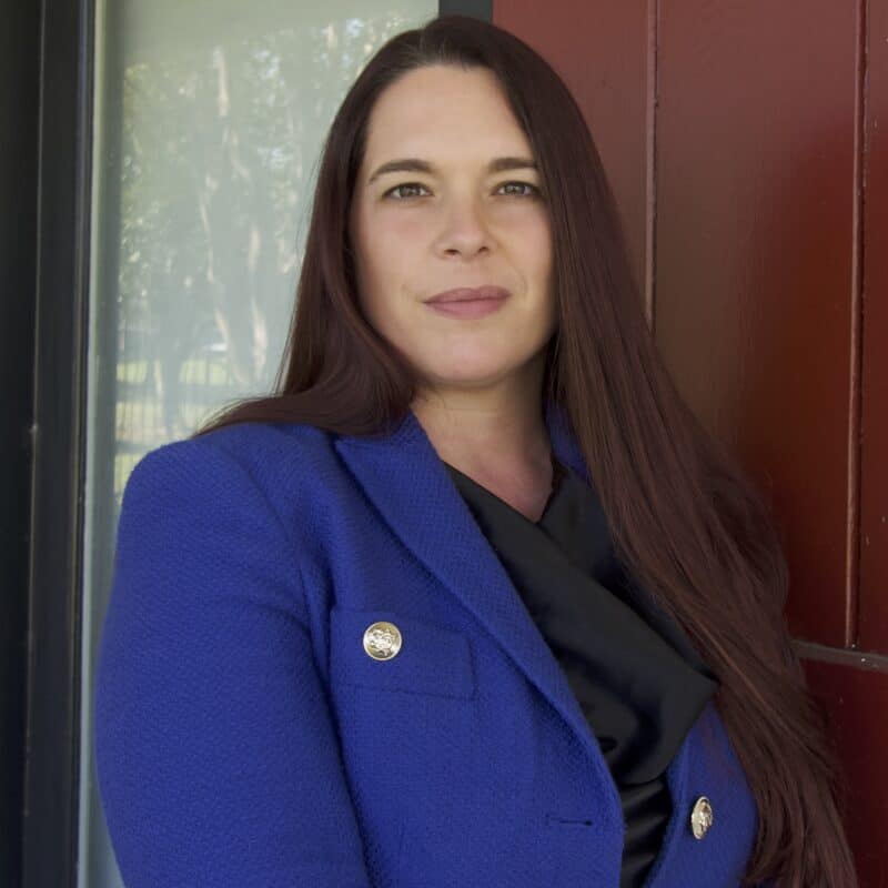 Woman with long brown hair wearing a blue blazer and black top stands in front of a red wall and window, looking at the camera.