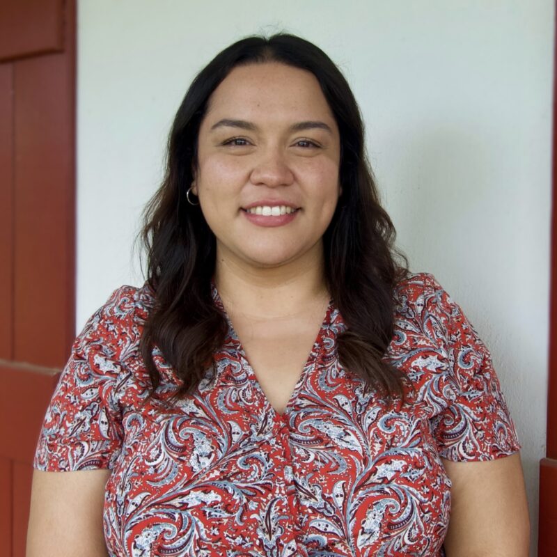 A woman with long, wavy dark hair smiles at the camera, wearing a red and white patterned top, in front of a white wall and red door.
