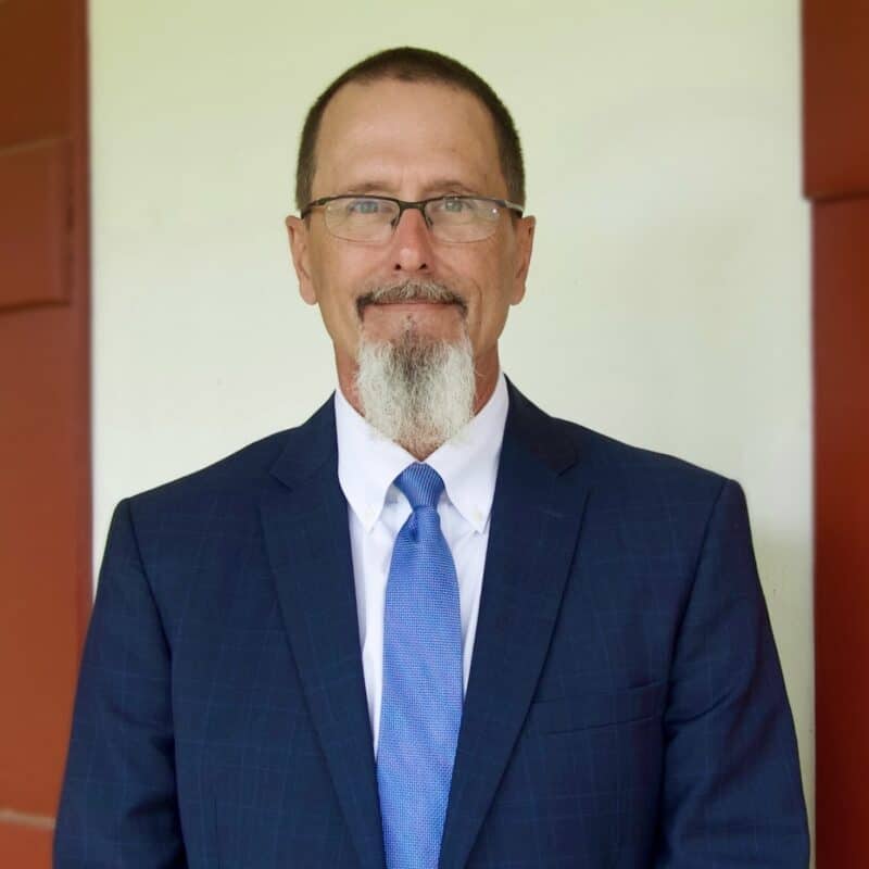 A man with glasses, a beard, and a mustache wears a blue suit and tie, standing in front of a cream wall with red trim.
