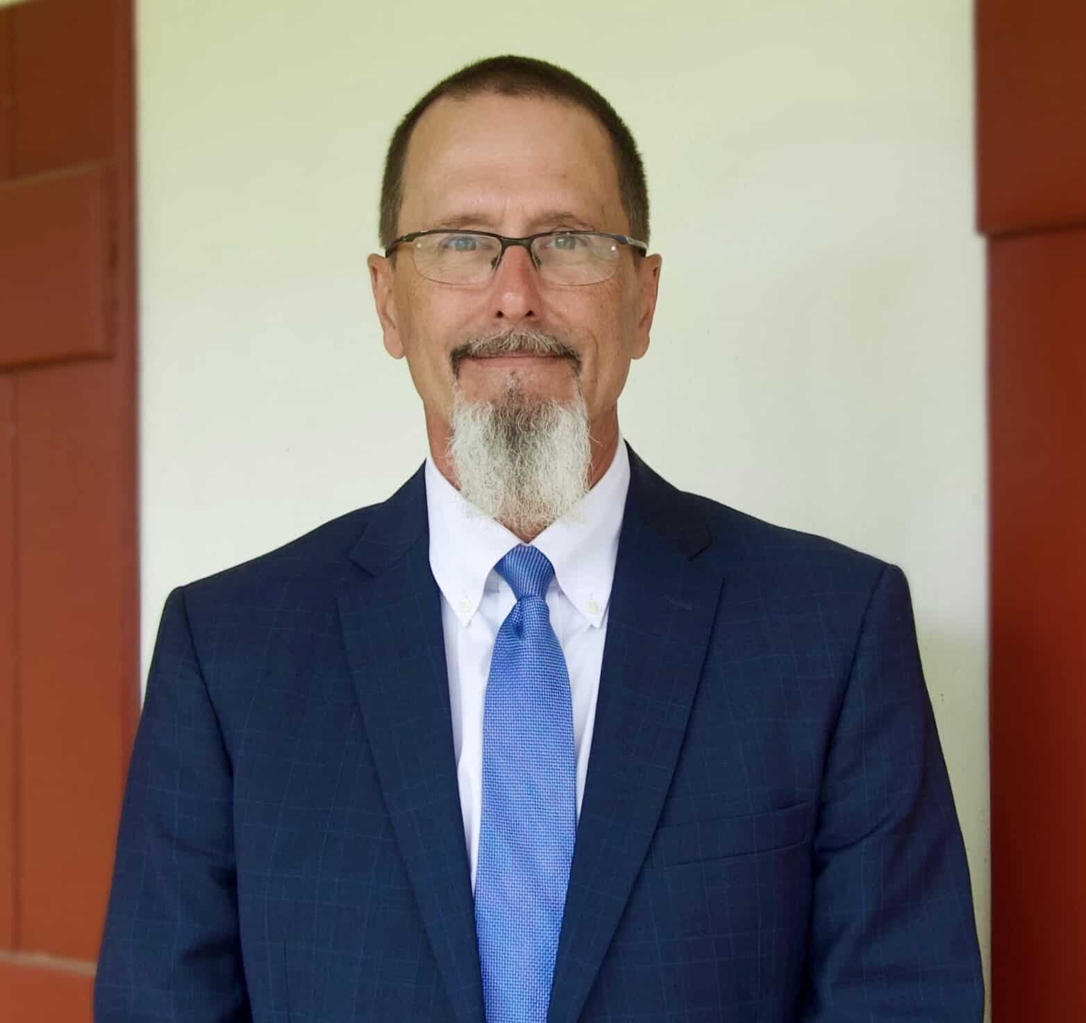 A man with glasses, a beard, and a mustache wears a blue suit and tie, standing in front of a cream wall with red trim.