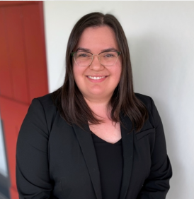 Woman with straight dark hair wearing glasses and a black blazer stands smiling in front of a white wall and red door.