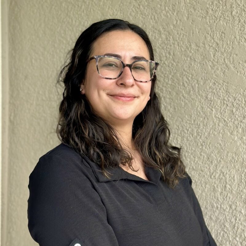 A woman with wavy dark hair and glasses stands in front of a textured beige wall, wearing a black collared shirt and smiling slightly.