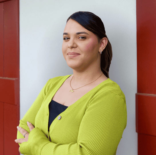 Woman in a lime green cardigan stands with arms crossed in front of a red and white wall, looking at the camera and smiling slightly.