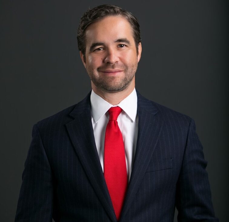 Man in a dark pinstripe suit, white shirt, and red tie stands against a plain dark background, looking at the camera and smiling.
