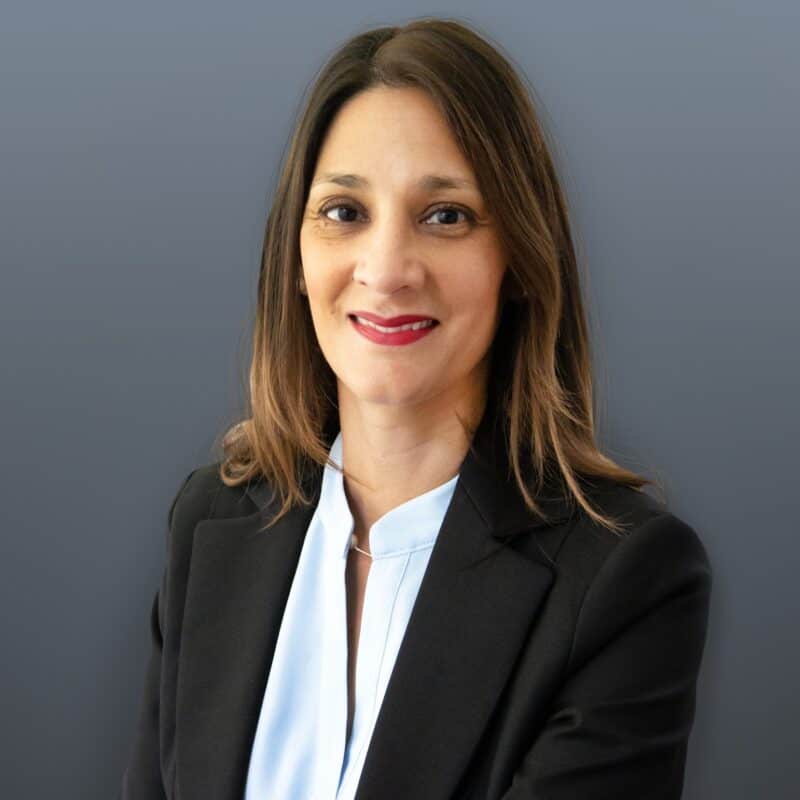 Woman with straight brown hair wearing a black blazer and light blue blouse, smiling in front of a plain gray background.
