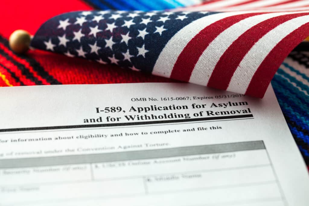 A close-up of an I-589 asylum application form with part of a folded U.S. flag, highlighting the topic of asylum filing fees.