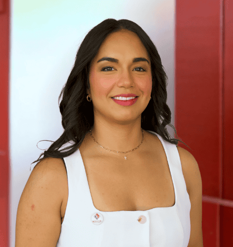 Woman with long dark hair in a white sleeveless top, smiling, standing in front of a red and white background.