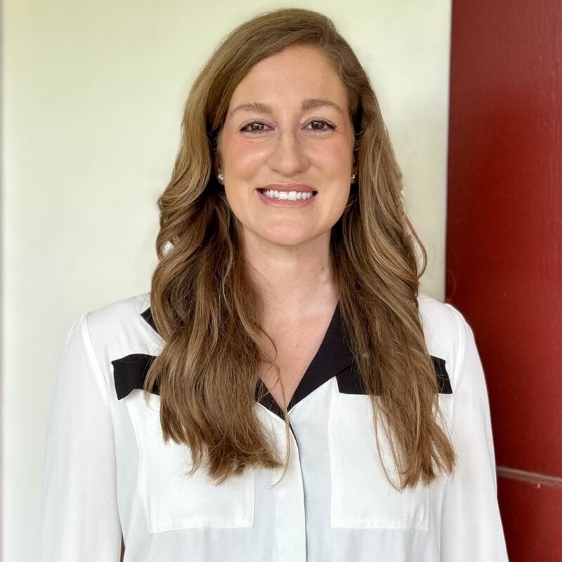 A woman with long brown hair wearing a white blouse with black accents stands in front of a white and red background, smiling.