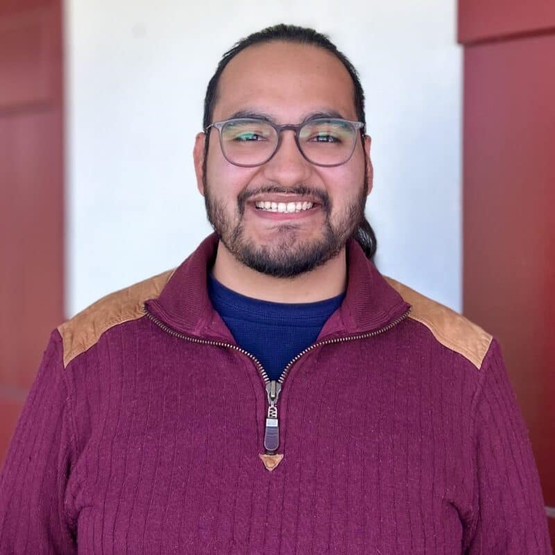 A person with glasses and a beard smiles, wearing a maroon sweater with tan patches, standing in front of a red and white background.